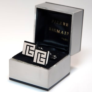 Puede incluir: Un par de gemelos de plata con un diseño geométrico en blanco y negro en una caja de plata con el texto "PIERRE BALMAIN PARIS" en la tapa.