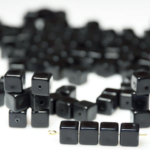 Puede incluir: Un montón de cuentas cuadradas negras con algunas apiladas unas sobre otras. Un hilo de cuentas cuadradas negras está en primer plano.