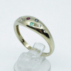 Może przedstawiać: Złoty pierścionek o kopulastym kształcie, osadzony serią małych kamieni szlachetnych. Kamienie szlachetne obejmują kamienie czerwone, zielone i przezroczyste. Pierścionek jest prezentowany na przezroczystym plastikowym stojaku.