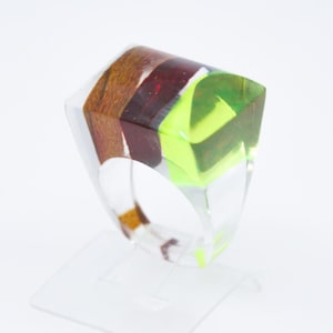 Könnte beinhalten: Ein klarer Harzring mit rechteckiger Oberseite. Die Oberseite weist Schichten aus Holz, rotem und grünem Harz auf. Der Ring wird auf einem transparenten Ständer präsentiert. Das Design des Rings ist modern und geometrisch.