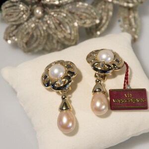 May include: A pair of gold-toned clip-on earrings with a floral design, featuring a central pearl and dangling teardrop-shaped pearl. The earrings have a tag that reads "vb voguebijoux". The earrings are displayed on a white pillow.