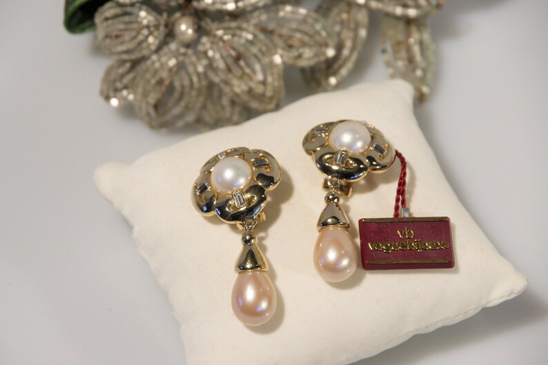 May include: A pair of gold-toned clip-on earrings with a floral design, featuring a central pearl and dangling teardrop-shaped pearl. The earrings have a tag that reads "vb voguebijoux". The earrings are displayed on a white pillow.