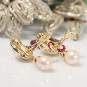 May include: A pair of gold-toned clip-on earrings with a pearl drop. The earrings have a decorative design on the clip and a small red accent. The background features a blurred, ornate, silver-colored floral arrangement.