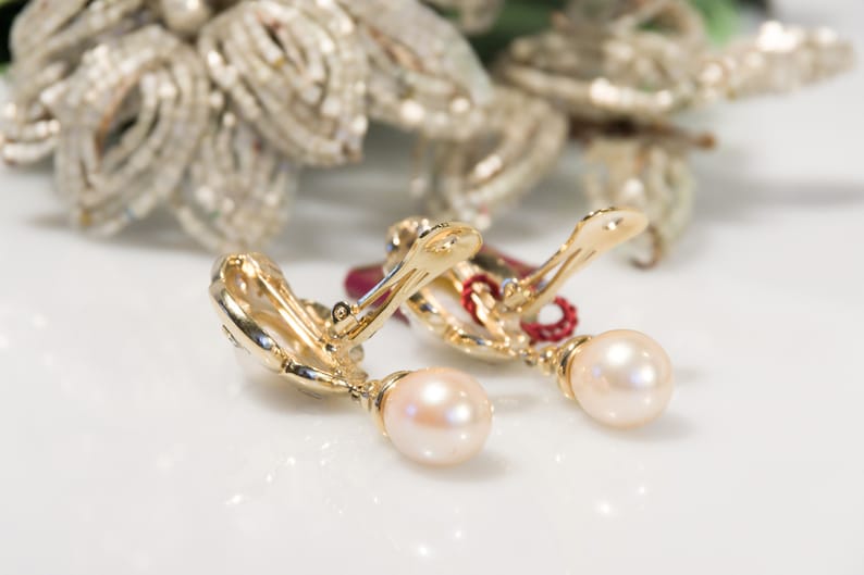 May include: A pair of gold-toned clip-on earrings with a pearl drop. The earrings have a decorative design on the clip and a small red accent. The background features a blurred, ornate, silver-colored floral arrangement.