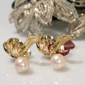 May include: A pair of gold-toned clip-on earrings with a pearl drop. The earrings have a floral design and a light pink pearl. A red string is attached to the earrings. The background features a decorative floral element.