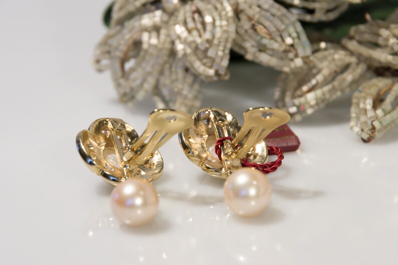 May include: A pair of gold-toned clip-on earrings with a pearl drop. The earrings have a floral design and a light pink pearl. A red string is attached to the earrings. The background features a decorative floral element.