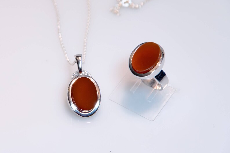 May include: A silver necklace and ring set with oval-shaped carnelian stones. The stones are a deep orange color and are set in silver.