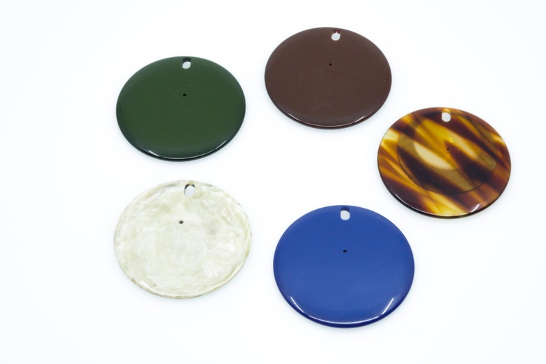 May include: Five round, flat, decorative ornaments with a hole in the center. The ornaments are in various colors, including green, brown, tortoiseshell, cream, and blue.