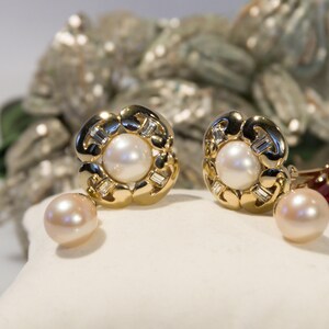 May include: Gold-tone earrings with a floral design, featuring a central pearl and surrounding baguette-cut stones. A matching pearl dangles below each earring. The earrings are displayed on a white surface, with a blurred background.
