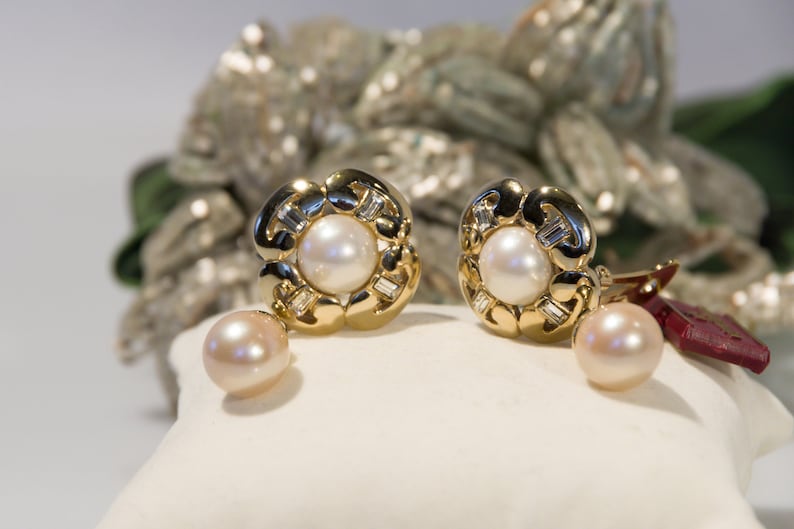 May include: Gold-tone earrings with a floral design, featuring a central pearl and surrounding baguette-cut stones. A matching pearl dangles below each earring. The earrings are displayed on a white surface, with a blurred background.
