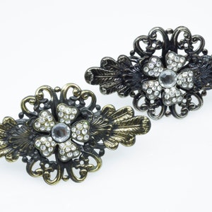 Könnte beinhalten: Zwei verzierte Haarklammern mit Blumenmuster. Jede Klammer hat eine große, klare Strassblume, die von kleineren Strasssteinen umgeben ist. Die Klammern sind aus dunklem Metall mit goldenen Akzenten.
