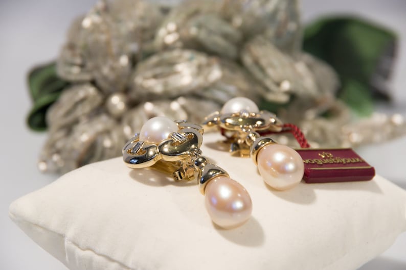May include: A pair of gold-toned earrings with a pearl accent. The earrings have a floral design with a large, round pearl at the top and a teardrop-shaped pearl hanging below. A red tag is attached.