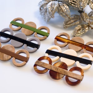 Könnte beinhalten: Vier Sätze Haarklammern mit verschiedenen Farbakzenten. Jede Klammer hat drei runde Teile, die durch eine Leiste verbunden sind. Die Farben sind Braun, Schwarz, Grau und Grün.