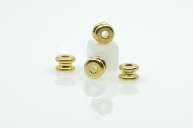 May include: Four gold-coloured metal beads with a round hole in the centre. The beads are stacked on top of each other, with the top bead resting on a white surface.