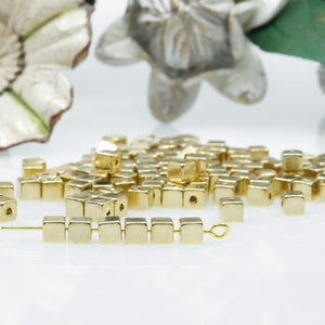 May include: A selection of small, gold-coloured square beads. Some beads are strung on a gold wire, while others are scattered. The beads have a metallic sheen and are evenly shaped. Decorative floral elements are in the background.