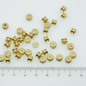 May include: A close-up of a group of gold-coloured metal beads. The beads are round with a hole in the centre. They are arranged in a random pattern on a white surface. A ruler is visible at the bottom of the image.