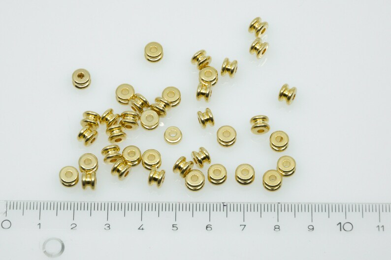 May include: A close-up of a group of gold-coloured metal beads. The beads are round with a hole in the centre. They are arranged in a random pattern on a white surface. A ruler is visible at the bottom of the image.
