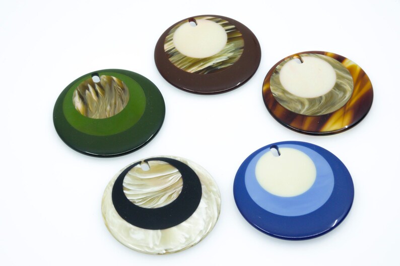 May include: Five round, decorative, resin pendants with a hole at the top for hanging. The pendants are layered with different colors and patterns, including brown, green, blue, black, and white.