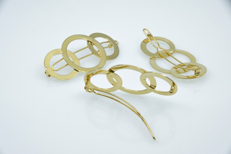 May include: Two gold hair clips. One clip is made of three interlocking circles. The other clip is made of five interlocking ovals.