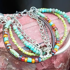 May include: A multi-strand bracelet featuring turquoise, yellow, and red beads, along with silver chains and a leather band. A silver heart charm and a flower charm dangle from the bracelet. The bracelet is displayed on a red surface.