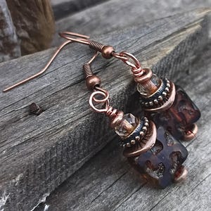 May include: A pair of dangle earrings featuring dark brown square glass beads with textured surfaces, accented by small clear faceted beads, antique copper-toned metal spacers, and copper wire-wrapped hooks for pierced ears.