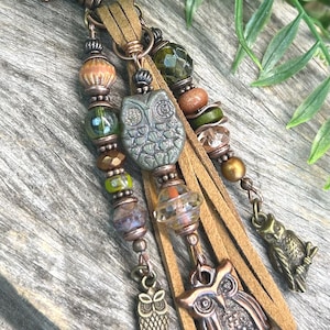 May include: A keychain featuring a copper-toned lobster clasp, tan suede leather strips, and multiple strands of beads in shades of green, brown, and clear. The design includes three owl-shaped charms in varying sizes and finishes, accented with metallic spacers and faceted glass beads.