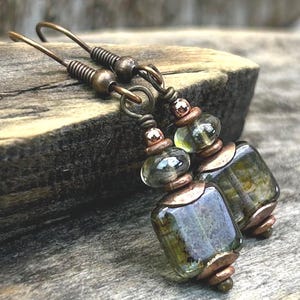 May include: A pair of dangle earrings featuring square, translucent olive-green glass beads accented with copper-toned metal spacers and findings. The earrings are suspended from copper-toned ear wires, showcasing a combination of smooth glass and metallic components.