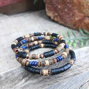 May include: A multi-strand wrap bracelet featuring a combination of cylindrical black, tan, and blue beads interspersed with small metallic copper-toned spacers. The design consists of several circular layers that coil around the wrist area.