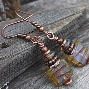 May include: A pair of dangle earrings featuring copper wire hooks and hardware. Each earring consists of a series of small copper beads and spacers alternating with translucent, square-shaped yellow glass beads that contain embedded copper wire accents.