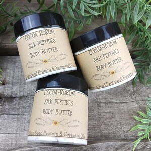 May include: Three jars of body butter with black lids and labels that read "COCOA-KOKUM SILK PEPTIDES BODY BUTTER" with "Hemp Seed Protein & Essential Oils" below. The jars are arranged on a wooden surface with green foliage.