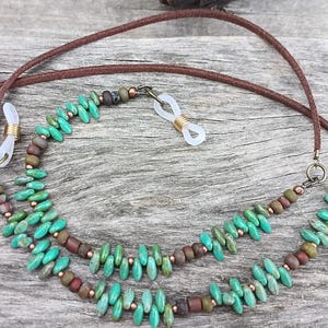 May include: A double-strand eyeglass chain featuring turquoise-colored beads in a rice shape, interspersed with small brown cylindrical beads and metallic copper accents. The chain includes brown leather cords and clear silicone loops with gold-tone coils.