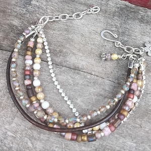 May include: A multi-strand bracelet featuring a mix of beads in various colors, including brown, pink, and gold. The bracelet includes a leather strand, a chain of rhinestones, and a silver-tone chain with a flower charm. The bracelet is secured with a hook clasp.