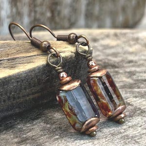 May include: A pair of dangle earrings featuring rectangular glass beads with a marbled pattern of brown, amber, and cream tones. The beads are accented with copper-toned metal caps and small beads, suspended from matching copper-toned ear wires.