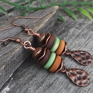 May include: Dangle earrings featuring copper-toned wire hooks and decorative caps. Each earring consists of three stacked circular beads in dark brown, light green, and tan, finished with a hammered, teardrop-shaped copper-toned metal charm.