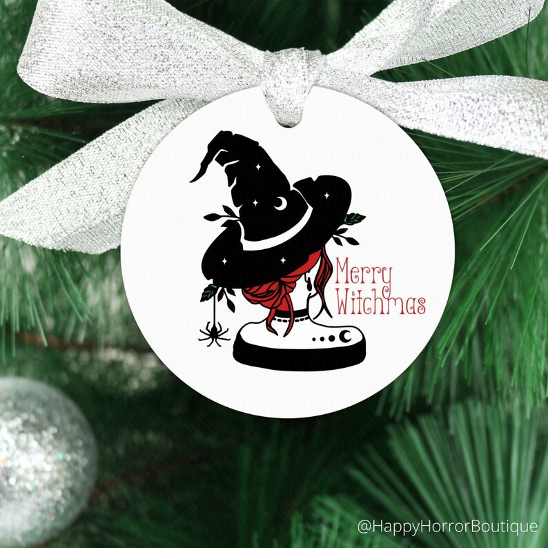 May include: A white round Christmas ornament with a black witch silhouette wearing a hat with stars and a crescent moon. The ornament has the text "Merry Witchmas" in red. A silver ribbon is attached to the ornament.