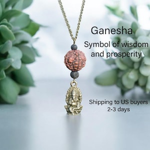 Puede incluir: Un collar de oro con un peque&ntilde;o colgante de Ganesha de lat&oacute;n y una cuenta de Rudraksha roja y marr&oacute;n. El collar est&aacute; sobre un fondo blanco con plantas verdes.