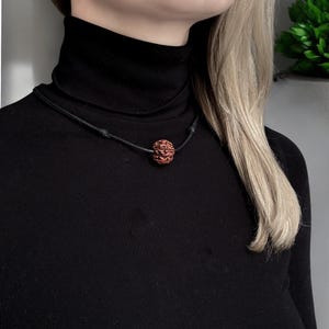 Könnte beinhalten: Eine Halskette mit einer einzelnen, runden, rotbraunen Rudraksha-Perle mit texturierter, gerillter Oberfläche. Die Perle ist mittig auf einem dünnen, glatten schwarzen Band platziert, das mit verstellbaren Knoten befestigt ist.