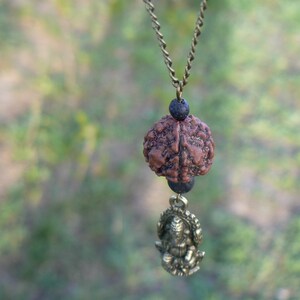 Puede incluir: Un collar marr&oacute;n y negro con un peque&ntilde;o colgante dorado detallado. El colgante es una representaci&oacute;n de una figura sentada. El collar est&aacute; hecho de una cadena con cuentas negras.