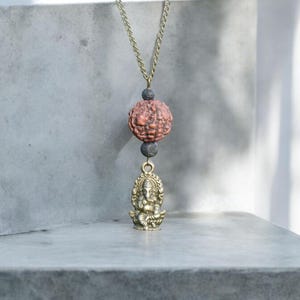 Puede incluir: Un collar de cadena dorada con una cuenta marr&oacute;n y negra y un colgante dorado de la deidad hind&uacute; Ganesha.
