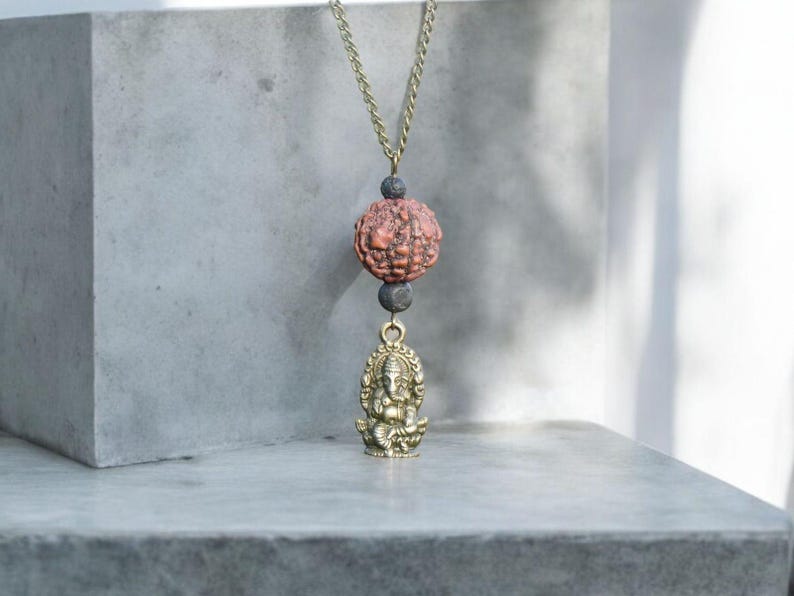 Puede incluir: Un collar de cadena dorada con una cuenta marr&oacute;n y negra y un colgante dorado de la deidad hind&uacute; Ganesha.