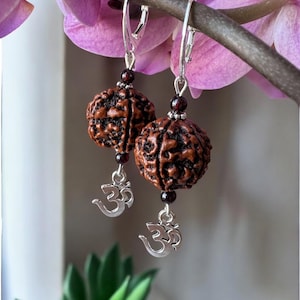 May include: A pair of silver earrings with brown wood beads and silver Om charms. The earrings are hanging from a branch with pink flowers.