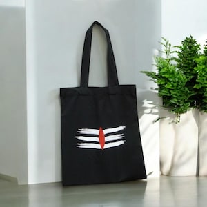 May include: A black tote bag with a white and red symbol printed on the front. The symbol is a stylized representation of the Hindu god Shiva's third eye.