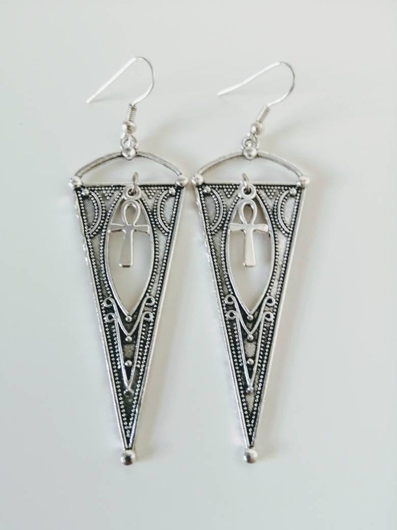 May include: Silver dangle earrings with a triangle shape and an ankh symbol in the center. The earrings have a detailed design with intricate patterns.