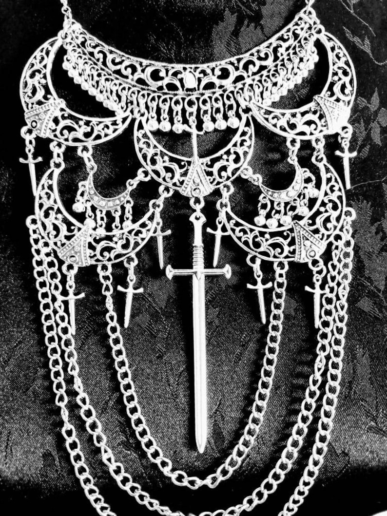 May include: A silver necklace with a large, ornate pendant featuring a sword design. The pendant is surrounded by smaller, crescent-shaped charms and hangs from a chain with multiple layers.