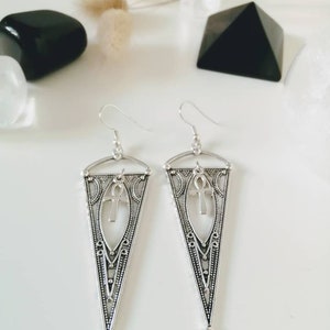 May include: Silver dangle earrings with a triangular design and an ankh symbol in the center. The earrings have a delicate, intricate pattern.