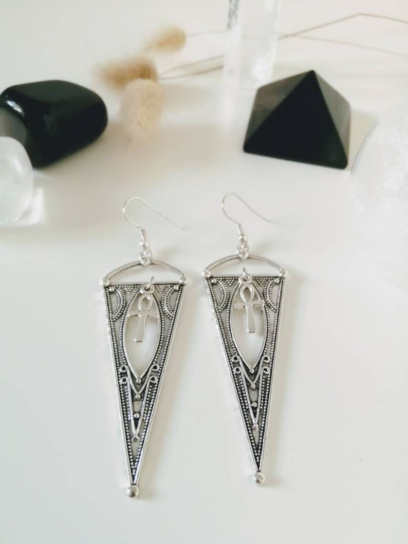 May include: Silver dangle earrings with a triangular design and an ankh symbol in the center. The earrings have a delicate, intricate pattern.