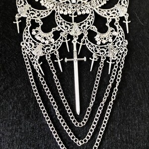 May include: A silver necklace with a large, ornate pendant featuring a sword design. The pendant is surrounded by smaller, intricate details and hangs from a chain with multiple strands.