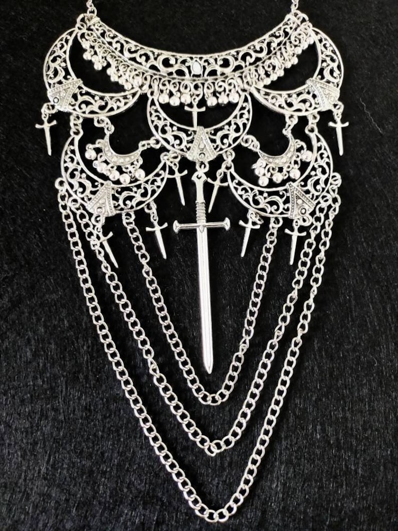 May include: A silver necklace with a large, ornate pendant featuring a sword design. The pendant is surrounded by smaller, intricate details and hangs from a chain with multiple strands.