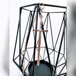Op de afbeelding: Een zilveren zwaardhangerketting die aan een zilveren ketting hangt. De hanger heeft de vorm van een zwaard met een puntige punt en een kruisbescherming. De ketting hangt aan een zwarte metalen standaard.