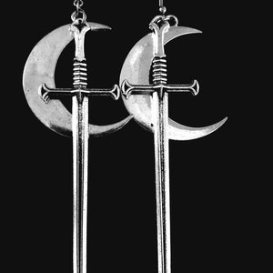 May include: Silver-toned earrings featuring a sword design suspended from a crescent moon. The earrings have a detailed sword with a cross-shaped hilt and a long, pointed blade. The crescent moon has a textured surface, and the earrings hang from a simple hook.